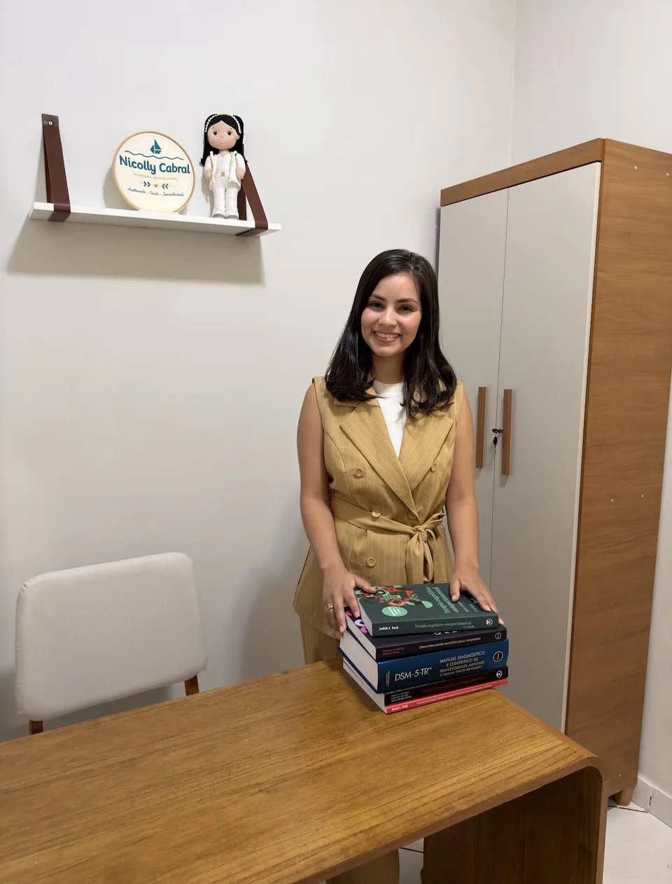 Nicolly no consultório, junto à mesa e aos livros de Psicologia